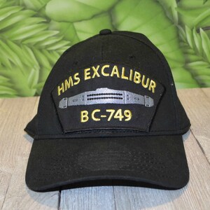 Könnte beinhalten: Schwarze Baseballkappe mit einem goldenen gestickten Aufnäher mit der Aufschrift "HMS EXCALIBUR BC-749".