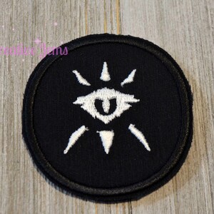 May include: A black circular patch with a white embroidered eye symbol. The eye has a white pupil and is surrounded by white rays.