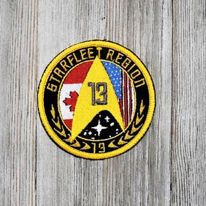 May include: A yellow and black embroidered patch with the text "Starfleet Region 13" and a Starfleet insignia. The patch features the Canadian and American flags.