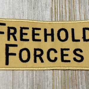 Peut inclure: Un écusson brodé de couleur beige avec des coutures noires qui indique "FREEHOLD FORCES".
