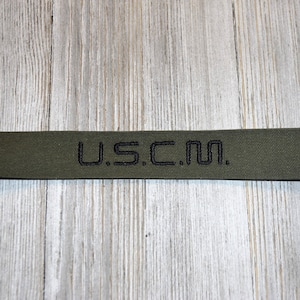 Może przedstawiać: Zbliżenie na zielony pasek z tkaniny z wyszytym czarnym tekstem "U.S.C.M.".