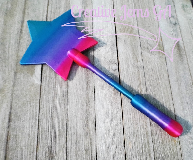 Peut inclure: Une baguette magique en forme d'&eacute;toile avec un d&eacute;grad&eacute; de bleu, de violet et de rose. La baguette a une longue poign&eacute;e et est pos&eacute;e sur un fond en bois. Le texte "Creative Jams Girl" est dans le coin sup&eacute;rieur droit.