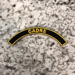 Può includere: Toppa nera e arcuata con la scritta "CADRE" ricamata in filo d'oro. La toppa ha un bordo dorato ed è posta su uno sfondo screziato grigio chiaro. La toppa è curva.