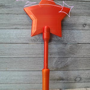 Peut inclure: Une baguette en forme d'&eacute;toile orange vif avec un long manche. La baguette pr&eacute;sente un motif d'&eacute;clat d'&eacute;toile et le texte "Creative Jems GA". La baguette est en plastique et est probablement un jouet ou un objet d&eacute;coratif.