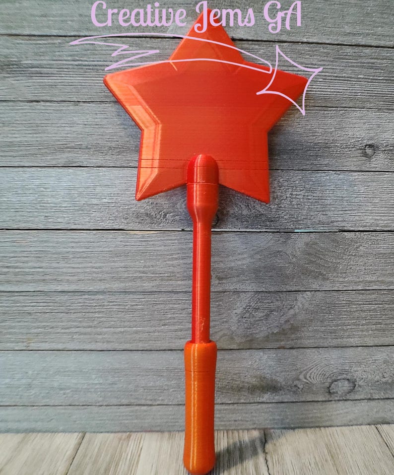 Peut inclure: Une baguette en forme d'&eacute;toile orange vif avec un long manche. La baguette pr&eacute;sente un motif d'&eacute;clat d'&eacute;toile et le texte "Creative Jems GA". La baguette est en plastique et est probablement un jouet ou un objet d&eacute;coratif.
