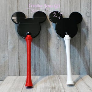 Peut inclure: Deux baguettes d&eacute;coratives noires en forme de Mickey Mouse. L'une a une poign&eacute;e rouge, l'autre une poign&eacute;e blanche. Elles sont pr&eacute;sent&eacute;es sur un fond en bois gris clair. Le texte "Creative Jems Gift" est visible en haut.