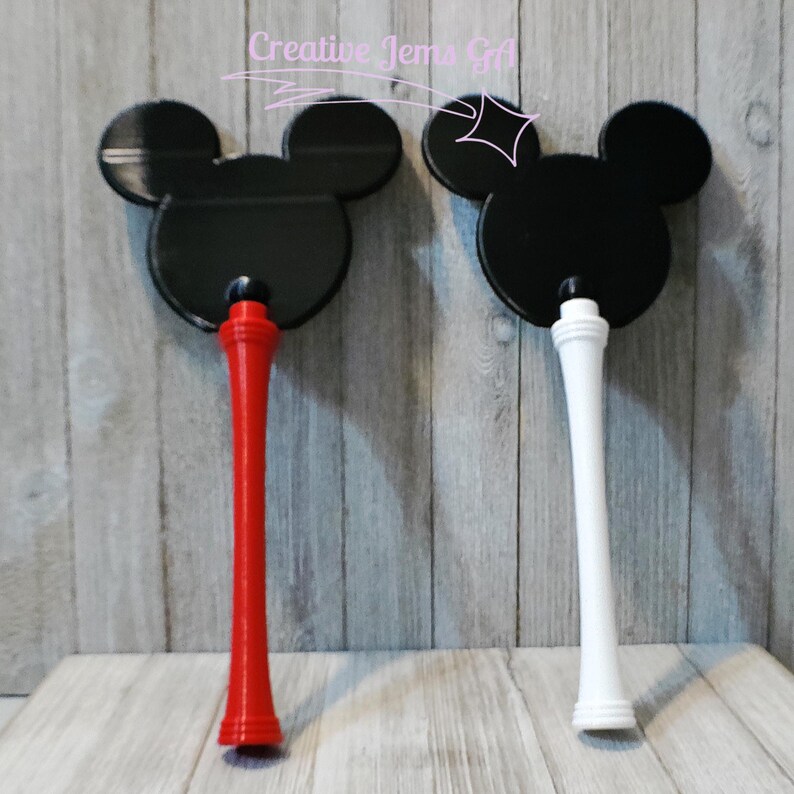 Peut inclure: Deux baguettes d&eacute;coratives noires en forme de Mickey Mouse. L'une a une poign&eacute;e rouge, l'autre une poign&eacute;e blanche. Elles sont pr&eacute;sent&eacute;es sur un fond en bois gris clair. Le texte "Creative Jems Gift" est visible en haut.