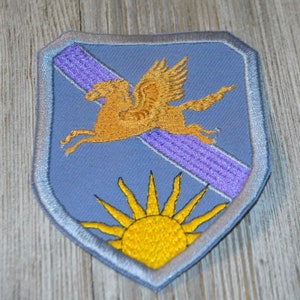 May include: A blue embroidered patch with a winged horse and a sun. The horse is gold and the sun is yellow.