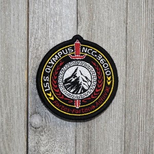 Puede incluir: Un parche bordado negro y amarillo con una montaña blanca y una espada. El texto en el parche dice "ISS OLYMPUS NCC-36010 Fortes Fortuna Adiuvat".