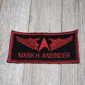 May include: Red embroidered patch with a black background. The patch features a red star with wings and the text "MARK H. ANBINDER".