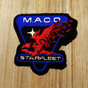 May include: Embroidered patch with the text "M.A.C.O." and "STARFLEET". The patch features a red eagle design on a black and blue background. The patch is rectangular with a black border.