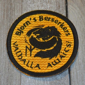 May include: A round, yellow embroidered patch with black stitching. The patch features a black dragon design with the text "Bjorn's Berserkers" and "Valhalla Aquatics" around the edge.