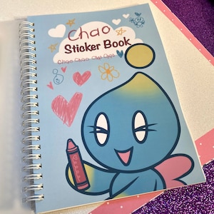 May include: A blue and yellow Chao character holding a red crayon on a blue spiral bound sticker book with the title "Chao Sticker Book" and the text "Chao Chao Chao Chao" in a white cloud.