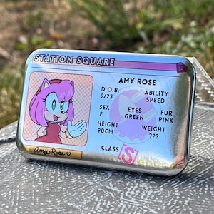 May include: A silver metal card featuring Amy Rose from Sonic the Hedgehog. The card includes Amy Rose's image, height (90cm), and other details. The text "STATION SQUARE" is at the top, and "Amy Rose" is written at the bottom.