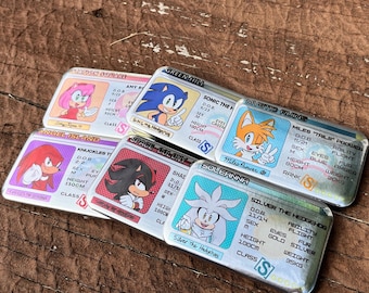 May include: A collection of rectangular metal cards featuring cartoon characters. Each card has a character illustration, name, and details like height (100cm or 110cm) and weight (20kg or 35kg). The cards are on a wooden surface.