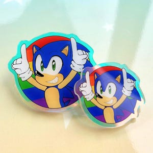 Op de afbeelding: Twee ronde acrylspeldjes met het personage Sonic the Hedgehog met blauwe vacht, beige huid en witte handschoenen, beide wijzend met beide handen omhoog. Elk personage staat tegen een levendige, veelkleurige ronde regenboogachtergrond.