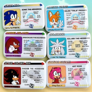 May include: Six rectangular pins featuring Sonic the Hedgehog characters: Sonic, Tails, Knuckles, Silver, Shadow, and Amy Rose. Each pin displays a character portrait, location name, and stats including D.O.B., ability, sex, eye color, fur color, height, and weight.