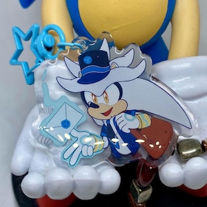 Könnte beinhalten: Ein transparenter Acryl-Anhänger mit einer Comicfigur mit weißem Fell, einem blauen Hut und einer blau-weißen Uniform. Die Figur hält einen weißen Umschlag mit einem blauen Siegel und eine braune Tasche. Der Anhänger ist an einem blauen sternförmigen Anhänger befestigt.