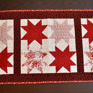 May include: A rectangular quilted table runner with a red and white star pattern. The runner features three white squares, each with a large red star and smaller stars in red and white floral patterns. The border is a dark red with small white dots.