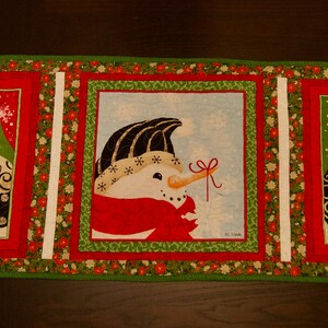 May include: A green, red, and white quilted table runner with three embroidered snowmen. Each snowman has a different colored hat and scarf. The snowmen are surrounded by a green border with red and white accents.