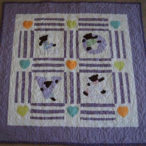 May include: A purple and white quilted baby blanket with a pattern of hearts and bears. The bears are wearing pajamas and the hearts are in various colors.