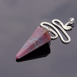 May include: A faceted pendulum with a maroon and teal speckled stone. The pendulum is suspended by a silver chain and a clear crystal bead. The object is set against a dark, reflective surface.