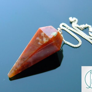 May include: A faceted, reddish-brown pendulum with a silver chain and a clear crystal bead. The pendulum has a pointed end and is likely used for divination or spiritual practices. The chain is attached to the top of the pendulum.