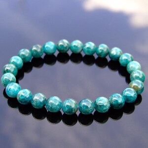 May include: A turquoise beaded bracelet. The round beads have a mottled appearance with shades of teal and green. The bracelet is displayed on a reflective surface, creating a mirrored effect. The beads are approximately 0.3 inches in diameter.
