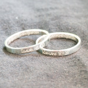 May include: Two silver rings with engraved bible verses. The top ring has the verse "Isaiah 41:10" and the bottom ring has the verse "Romans 8:37".