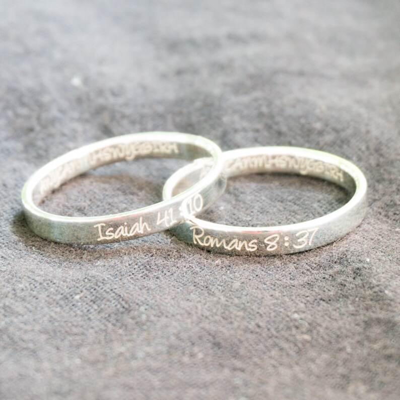 May include: Two silver rings with engraved bible verses. The top ring has the verse "Isaiah 41:10" and the bottom ring has the verse "Romans 8:37".