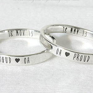 May include: Two silver rings with engraved text. One ring reads "PROUD" with a heart symbol and "OH", the other reads "OH" with a heart symbol and "PROUD". Both rings have "18 NOV 13" engraved on them.