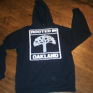Puede incluir: Sudadera con capucha negra con un gráfico blanco de un árbol y el texto "Rooted in Oakland".