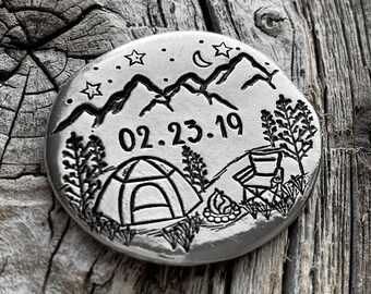 May include: A silver round charm with an engraved design of a mountain range, a tent, a campfire, and stars. The date 02.23.19 is engraved on the charm.