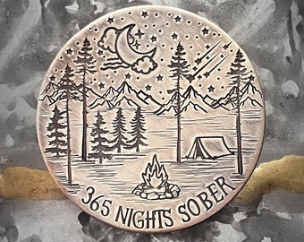 May include: A round, copper-colored coin with a design of a night sky with a crescent moon, stars, and a shooting star. The coin also features a campfire, a tent, and trees. The text "365 Nights Sober" is inscribed around the edge of the coin.
