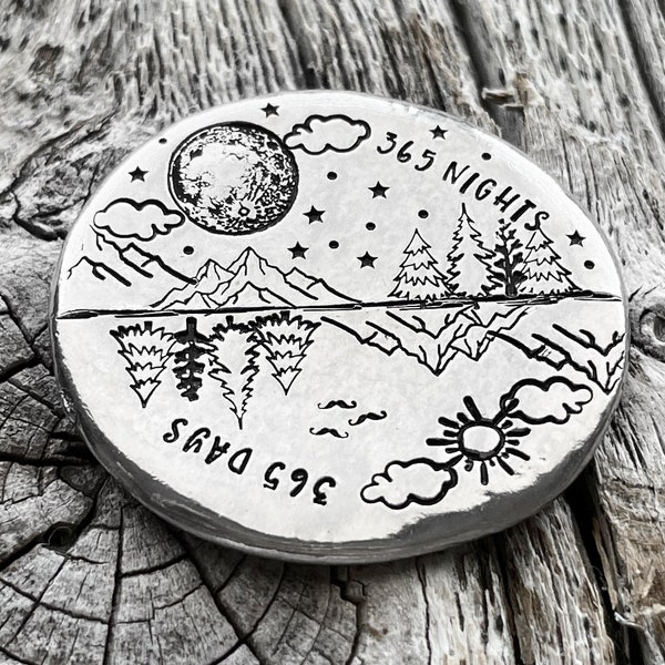 May include: A silver round disc with an engraved design of a moon, stars, mountains, trees, and a sun. The text "365 Days" and "365 Nights" is engraved on the disc.