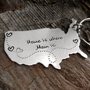 Puede incluir: Un llavero de plata con forma de Estados Unidos con el texto "Home is where Mom is" y una l&iacute;nea punteada con corazones a lo largo del borde.