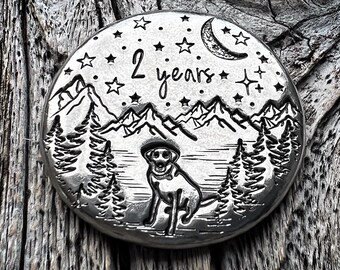May include: A silver round disc with an engraved image of a dog standing in front of a mountain range with a crescent moon and stars in the sky. The text "2 years" is engraved on the disc.