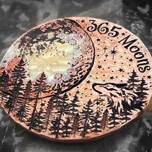 May include: A copper coin with a detailed design of a crescent moon, a wolf howling at the moon, and a forest of trees. The coin is embossed with the text "365 Moons".