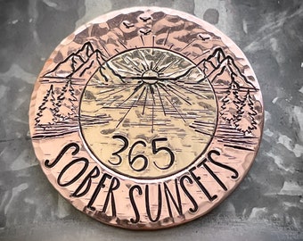 May include: A copper-colored round metal medallion with the words "SOBER SUNSETS" and the number "365" engraved on it. The center features a mountain and sun design. The background is a textured, metallic surface.