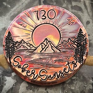 May include: A round, copper-colored metal item with a sunset design. The image features a sun, mountains, trees, and the text "730" and "Sober Sunsets". The item has a rustic, engraved appearance.