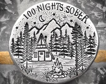 May include: A silver coin with the text "100 Nights Sober" engraved around the edge. The coin features a cabin in the woods with a campfire in front of it. There are stars and a crescent moon in the sky.