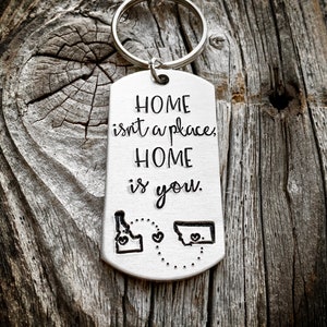 以下が含まれることがあります： 「Home isn't a place, Home is you.」というテキストと、ハートマークが付いたアイダホ州とモンタナ州の輪郭が描かれたシルバーのキーホルダーです。
