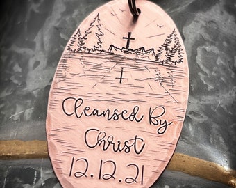 Könnte beinhalten: Ein ovaler Schlüsselanhänger aus Kupfer mit einer eingravierten Kreuz- und Berglandschaft. Der Text "Cleansed By Christ 12.12.21" ist unten eingraviert.