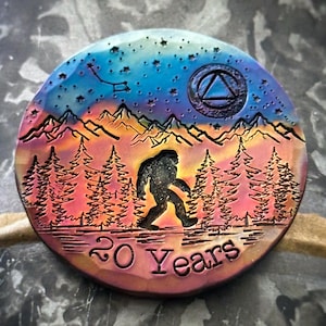 May include: A round, metallic coin with a colorful, iridescent finish. The coin features a silhouette of Bigfoot walking through a forest with mountains in the background. The sky is a vibrant blue with stars and a crescent moon. The text "20 Years" is engraved at the bottom of the coin.
