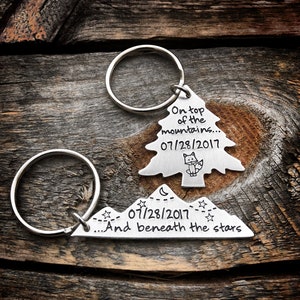 May include: Two silver keychains with a mountain and tree design. The mountain keychain says "...And beneath the stars" and "07/28/2017". The tree keychain says "On top of the mountains..." and "07/28/2017".