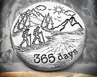 May include: A silver round disc with an engraved image of two hikers walking through a mountain landscape with a sun and clouds in the sky. The text "365 days" is engraved below the image.