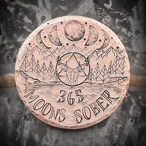 May include: A copper-colored round coin with an engraved design. The design features a deer head inside a triangle, surrounded by trees and a crescent moon. The text "365 Moons Sober" is engraved around the edge.