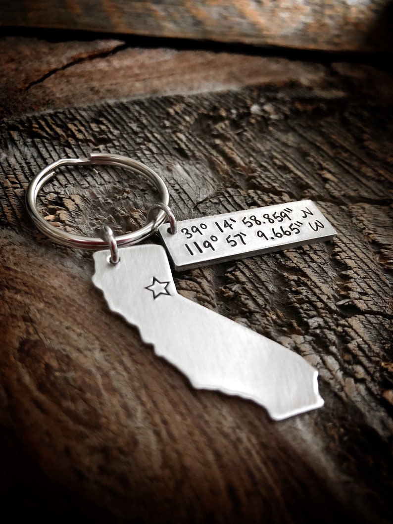 May include: Silver keychain featuring a California state outline with a star inside and a tag with the coordinates 39&deg; 14' 58.859" N 119&deg; 57' 9.665" W.