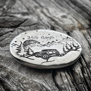 May include: A silver round coin with an engraved design of a jeep driving through a mountain landscape with a full moon and stars. The text "365 days" is engraved above the jeep.