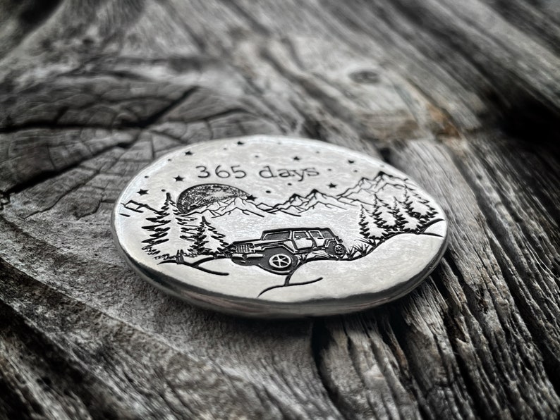May include: A silver round coin with an engraved design of a jeep driving through a mountain landscape with a full moon and stars. The text "365 days" is engraved above the jeep.